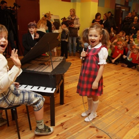 Od lewej chłopiec udający grę na makiecie fortepianu, obok śpiewająca dziewczynka, w tle dzieci uczestniczące w imprezie siedzą na podłodze w Centrum Kultury Dziecka