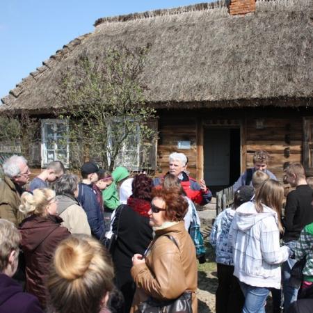 Uczestnicy wycieczki słuchają przewodnika stojącego przed chatą w skansenie